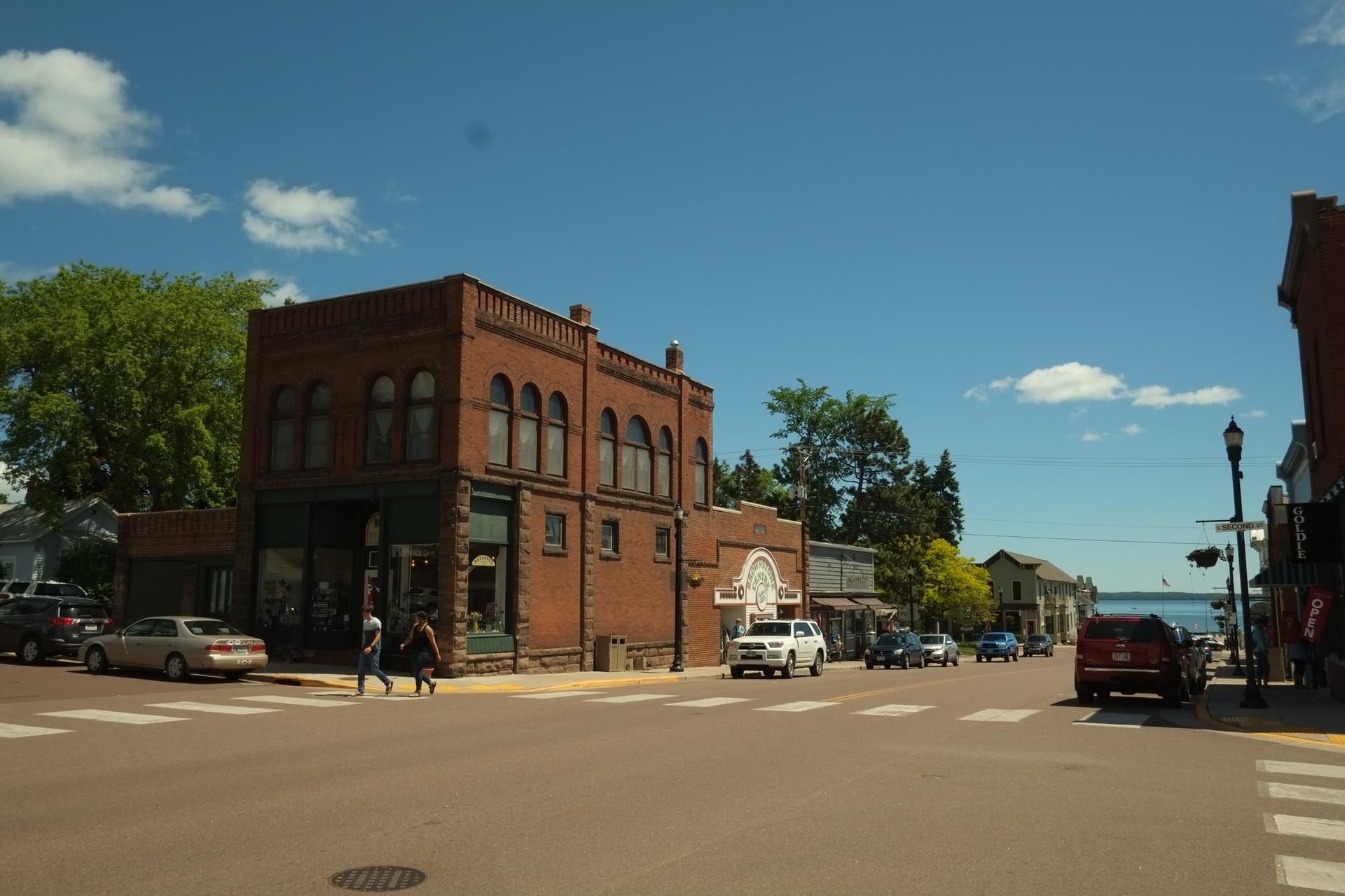 Sunny street scene with brick buildings, cars, and people walking.