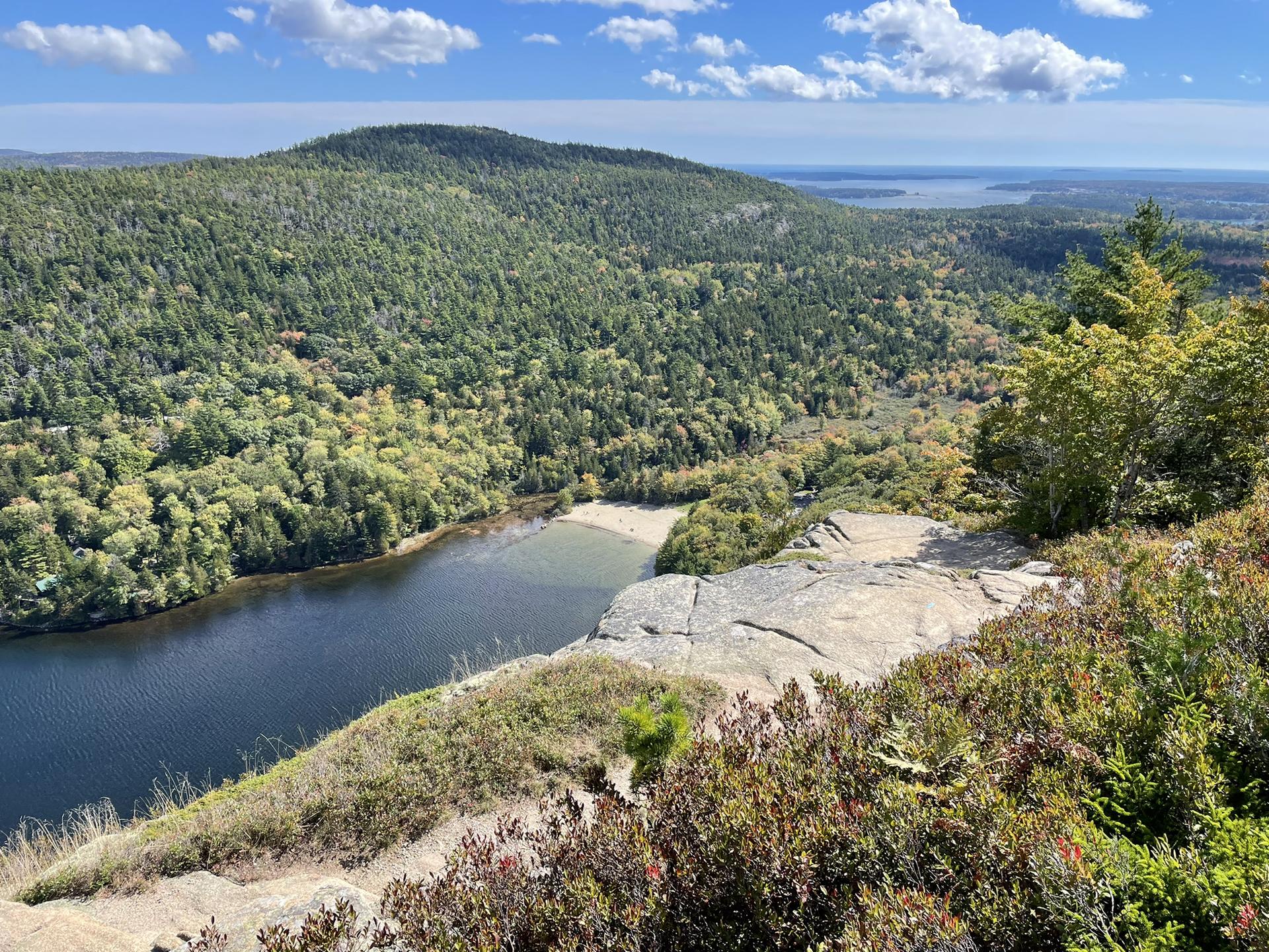 Overlook of a lake and sandy shore surrounded by green forested hills under a blue sky.