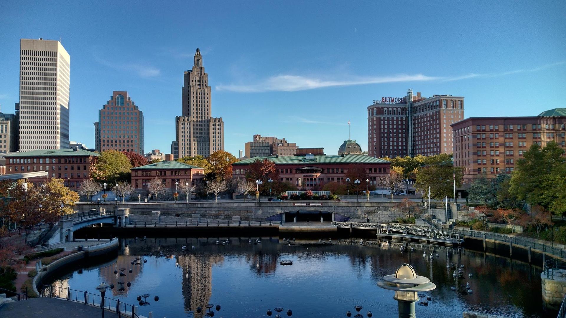 City skyline with tall buildings reflecting in calm water on a clear day.