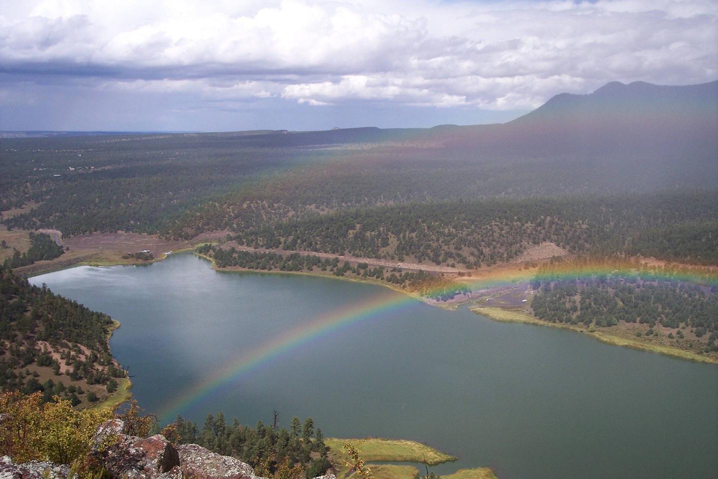 A scenic lake and forest under a cloudy sky with a vibrant rainbow.