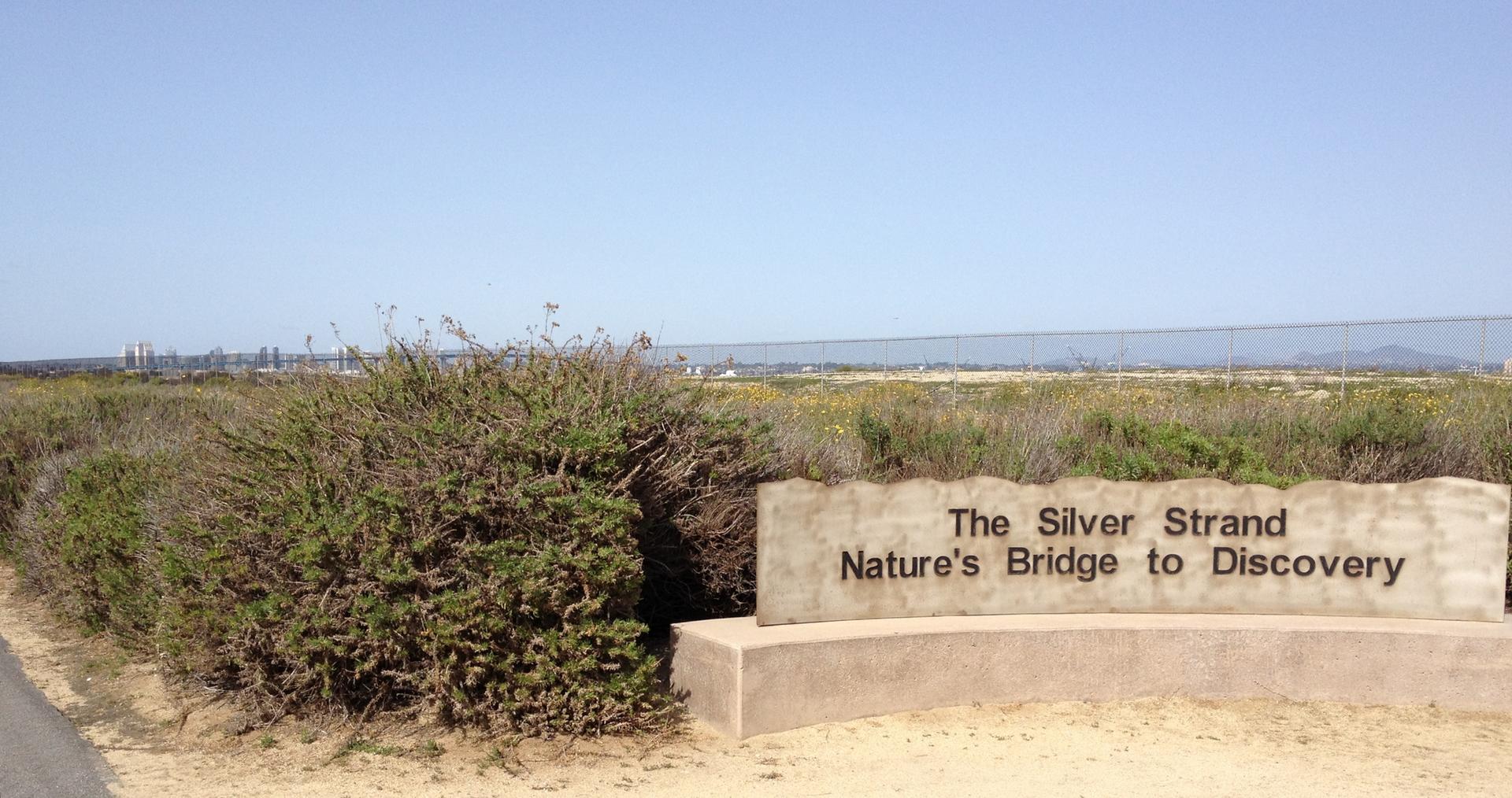 Stone sign in front of green shrubs, with distant industrial structures.