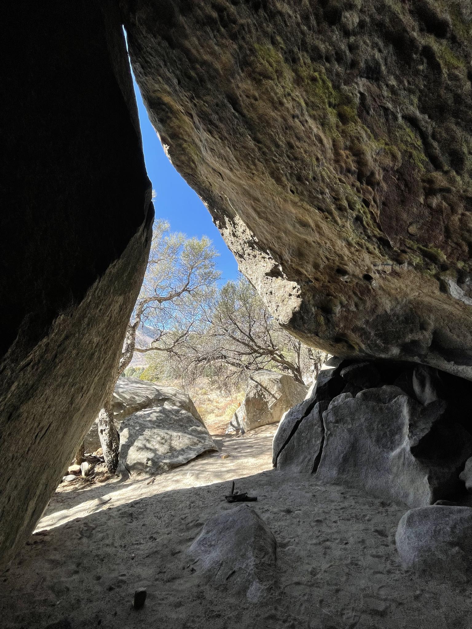 View from a dark rock cave opening to sunlit trees and blue sky.