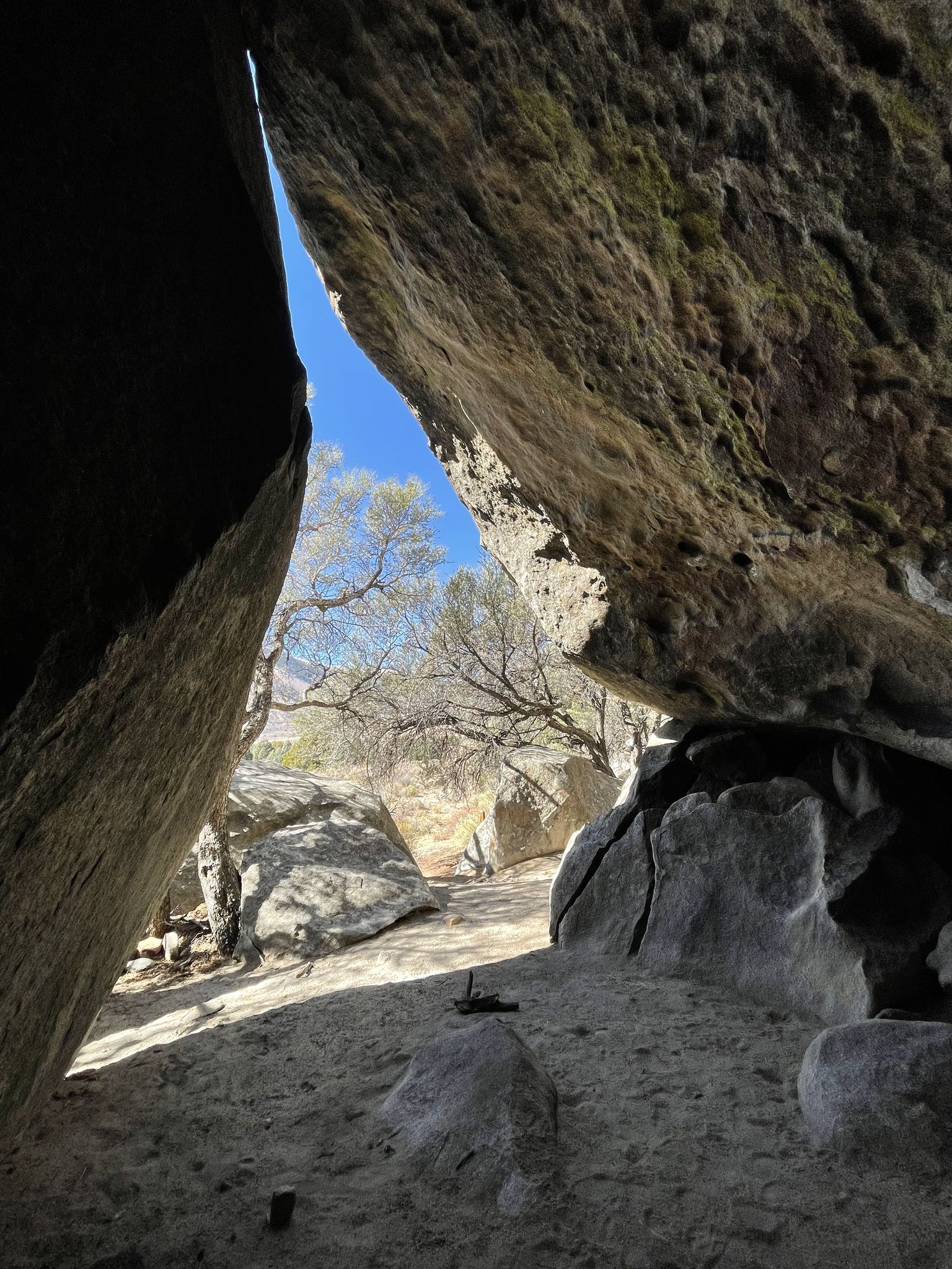 View from a dark rock cave opening to sunlit trees and blue sky. - Backyard Boulders Trail Loop