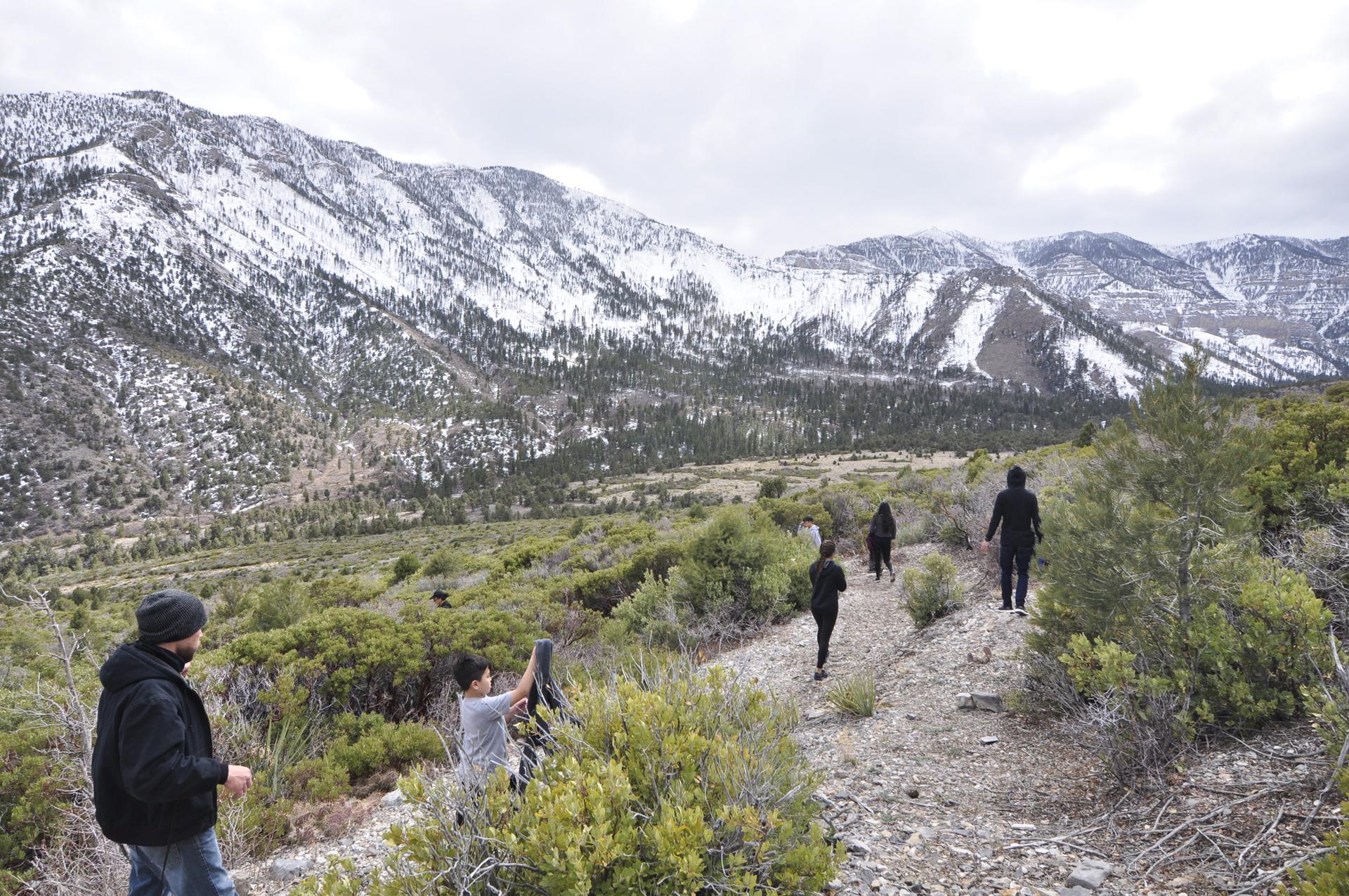 Hikers on a dirt path with overcast skies and snow-capped mountains.