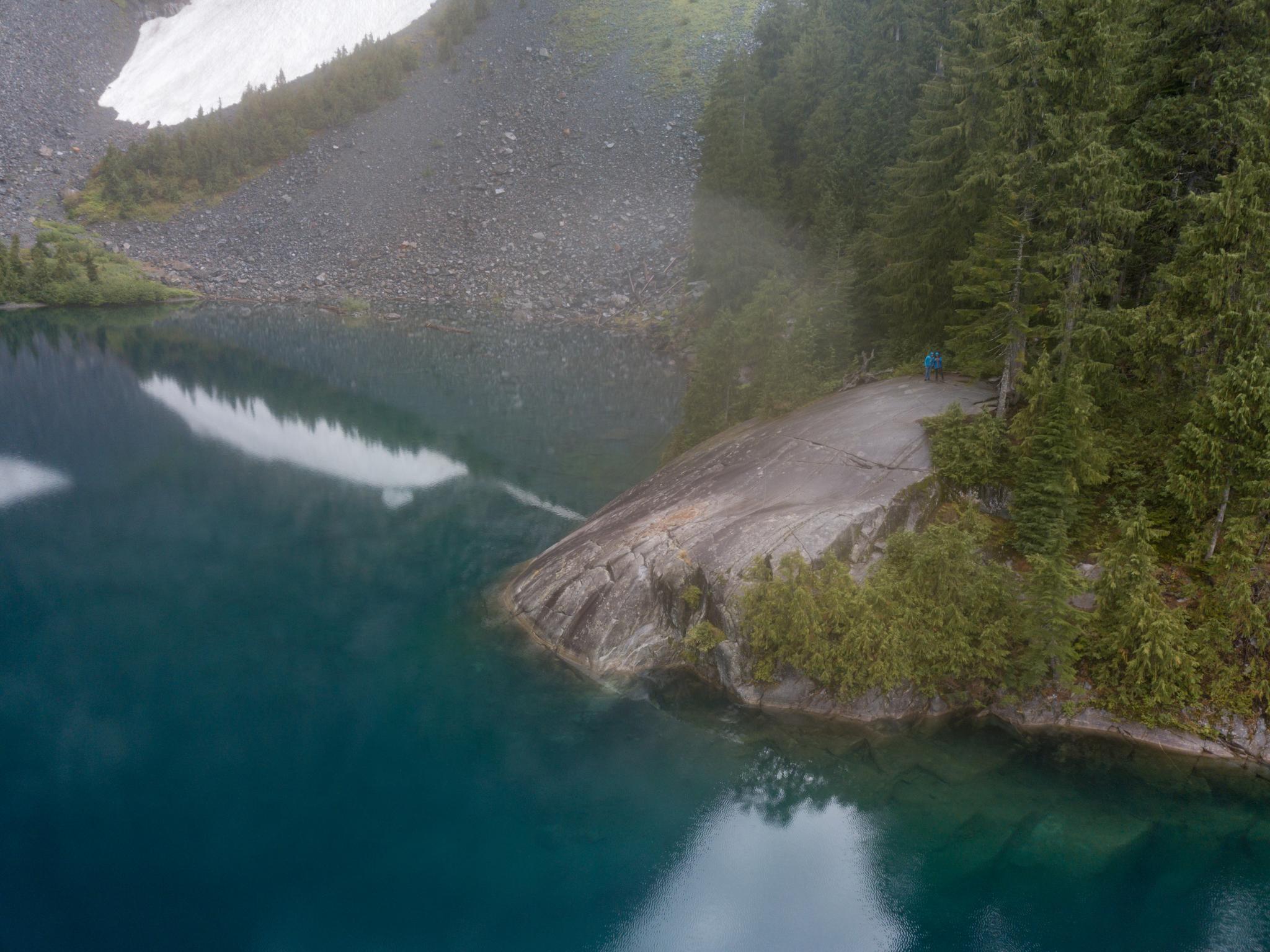 A person sits on a rocky outcrop by a clear turquoise mountain lake.