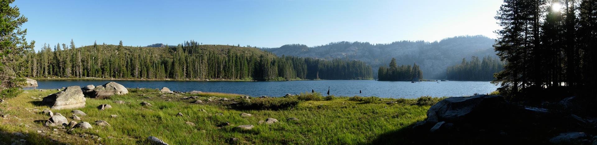 Panoramic view of a calm lake surrounded by dense forests under a bright sky.