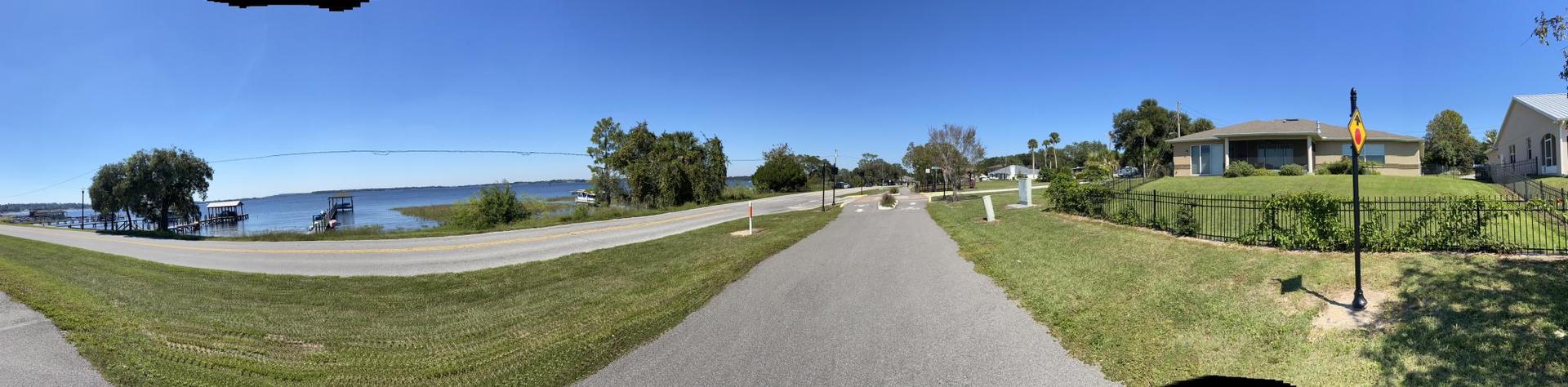 Panoramic view of a paved path leading to a sunny waterfront with houses.