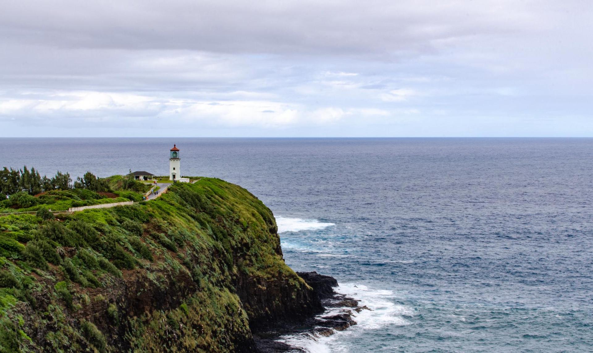 White lighthouse on a green cliff overlooking a vast, wavy ocean under cloudy skies.