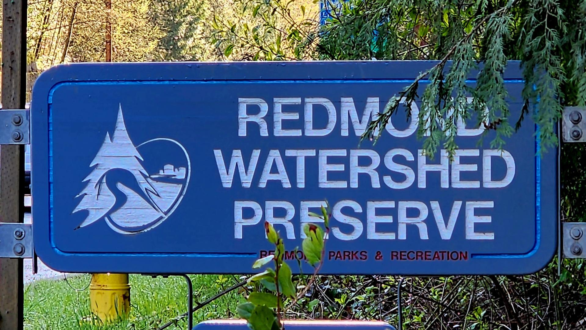 A blue sign with white text and a nature logo, surrounded by green foliage.