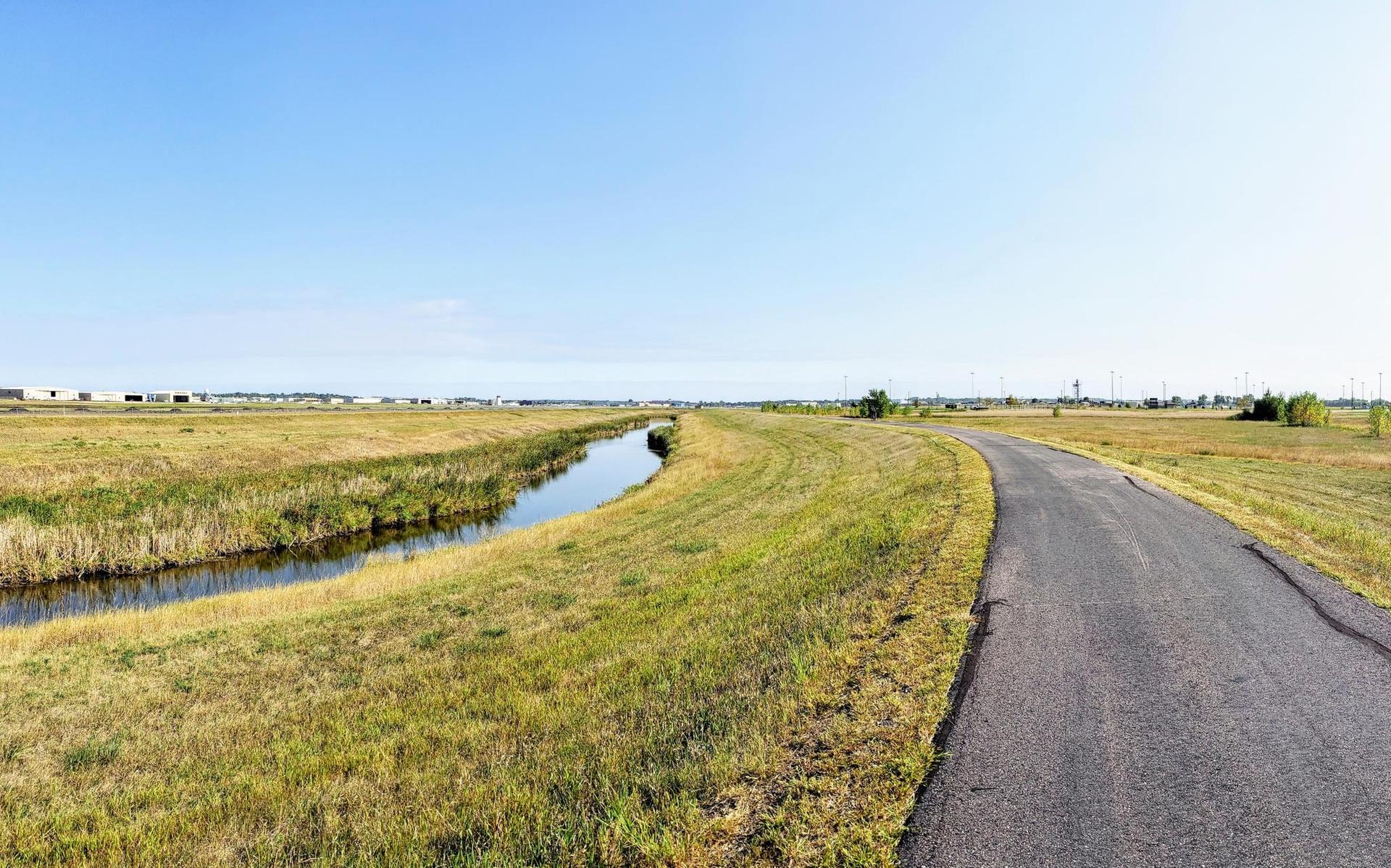 Paved path next to a narrow canal through flat grassy fields under clear sky.