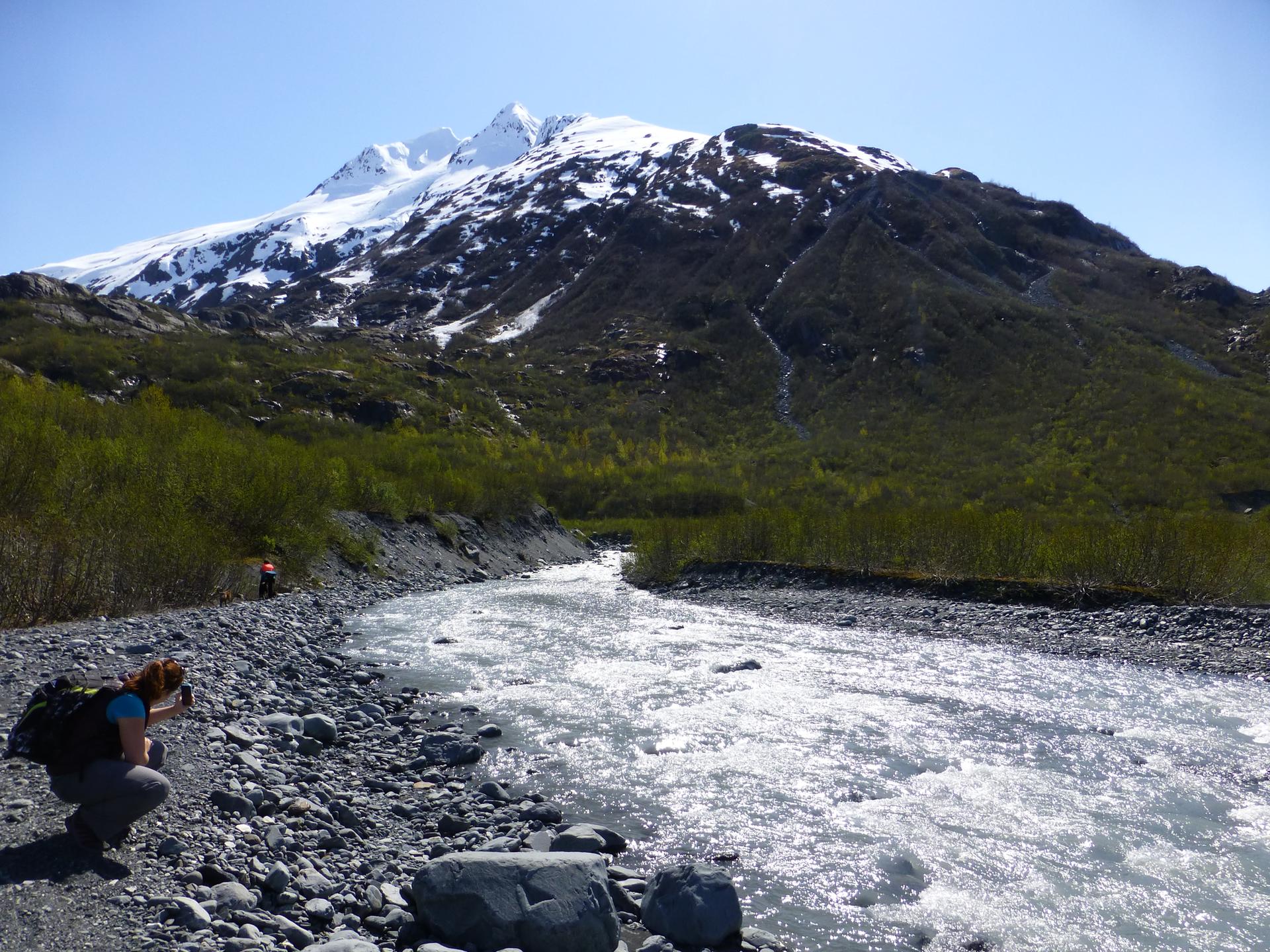 A person crouches by a rocky river, with a snow-capped mountain and green trees.