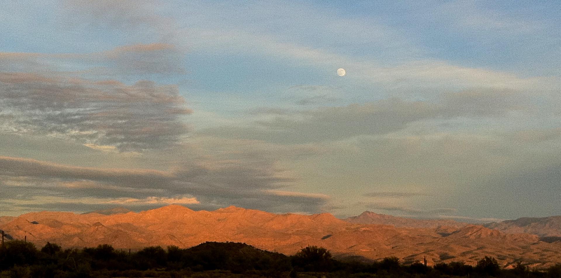 Golden mountains under a cloudy sky with a visible moon at dusk.