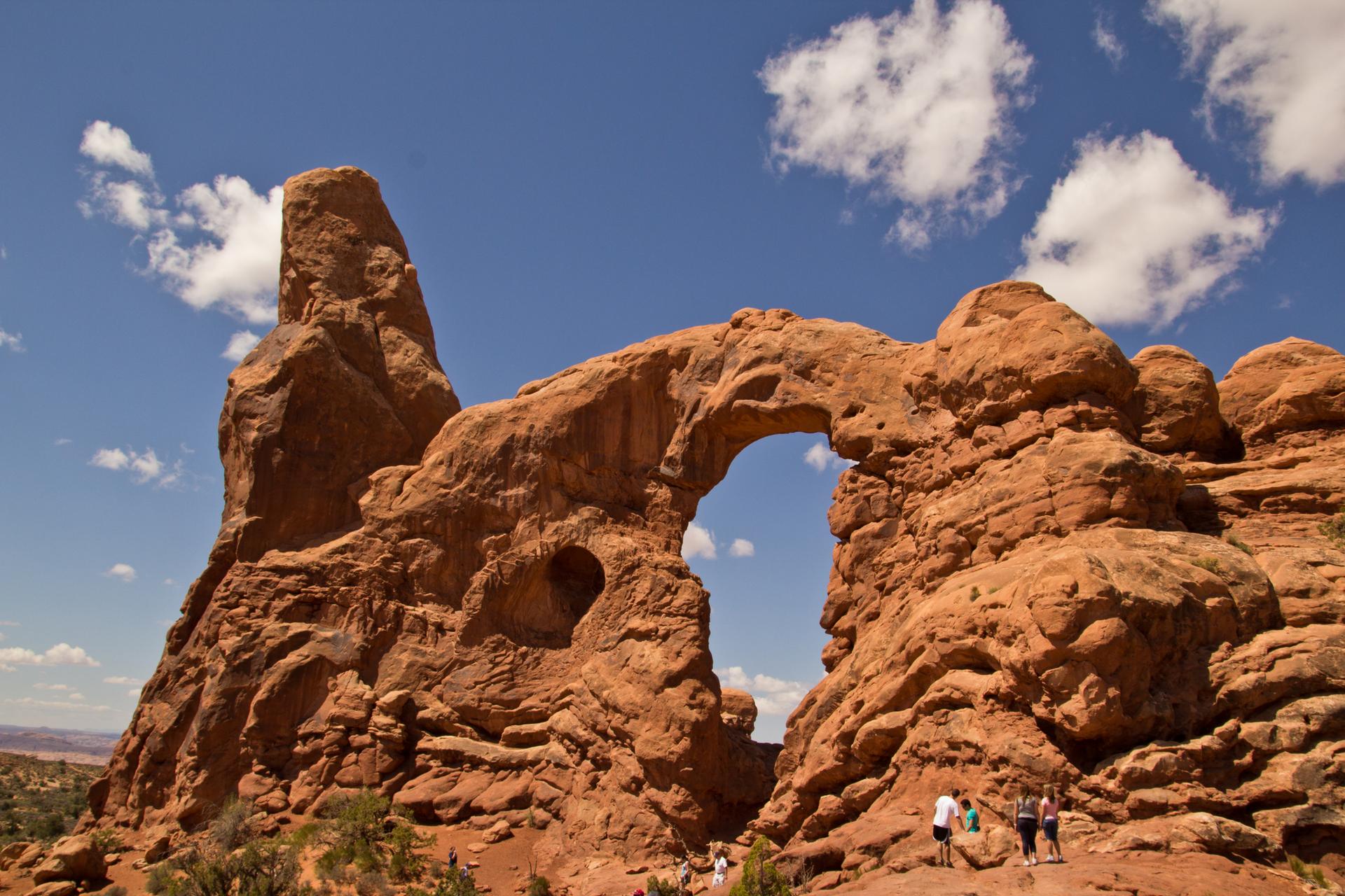 Tall red rock arch with a pointed formation, people walking below.