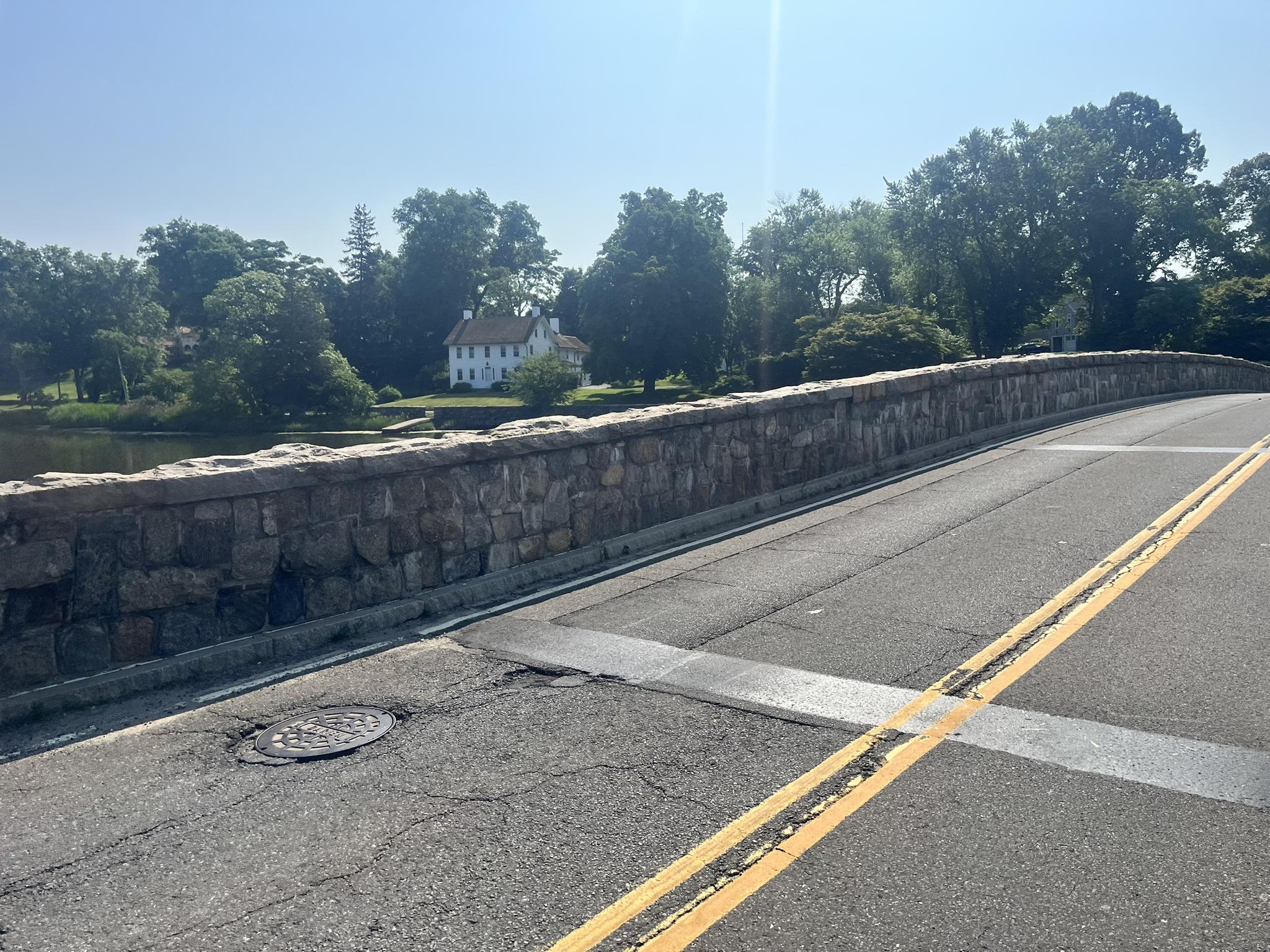 Stone bridge with a road, trees, and a distant white house.