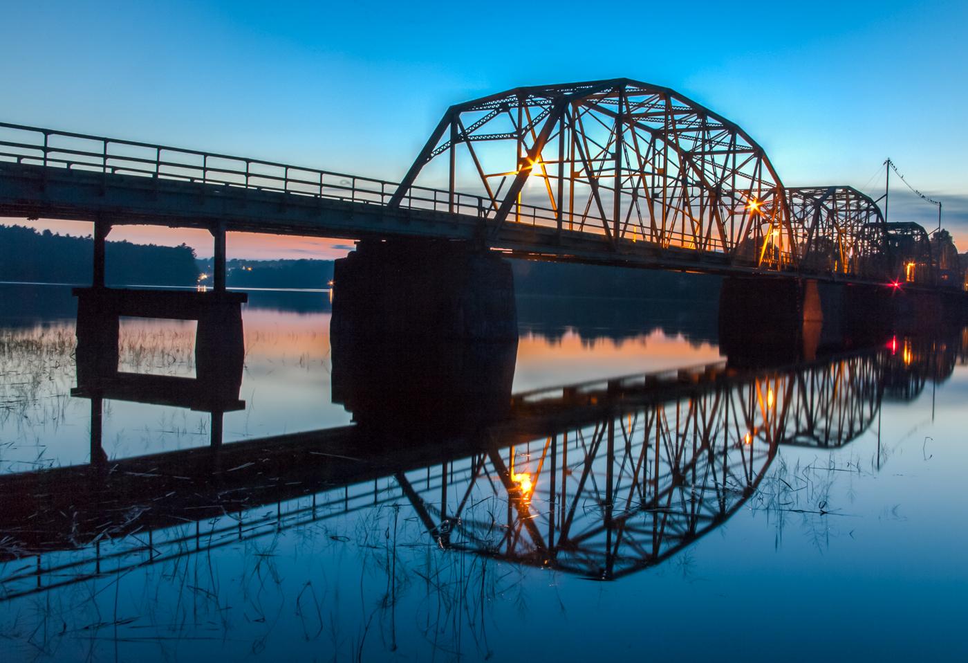 Illuminated metal truss bridge reflected in calm water at twilight.