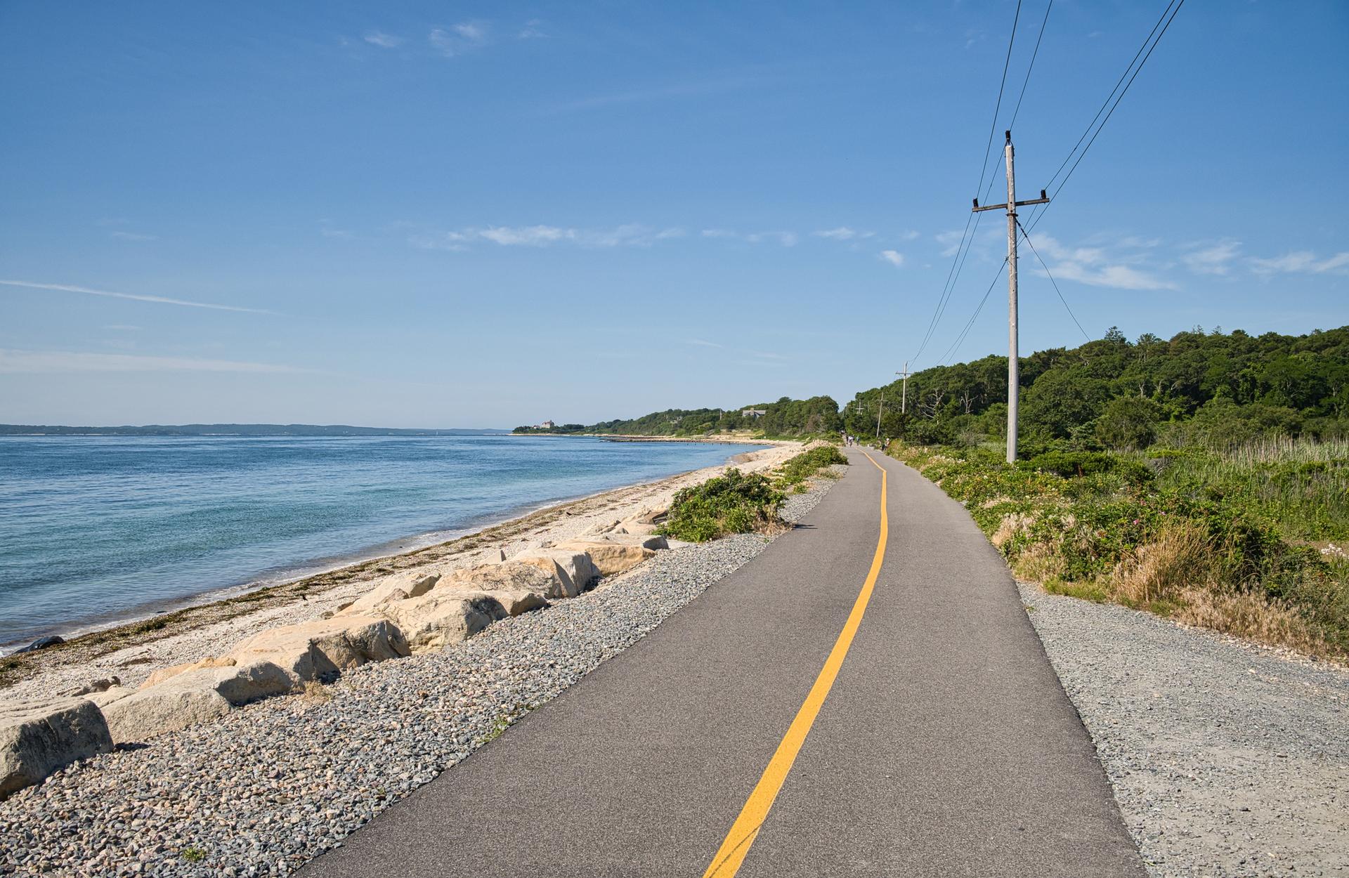 Paved path with yellow line runs alongside a sunny beach and blue ocean.
