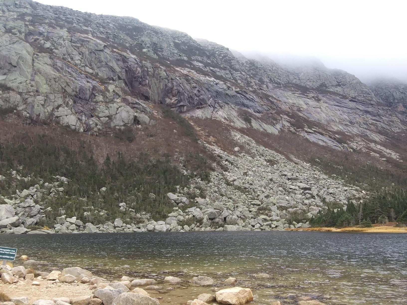 Dark lake at the base of a steep, rocky mountain under an overcast sky.