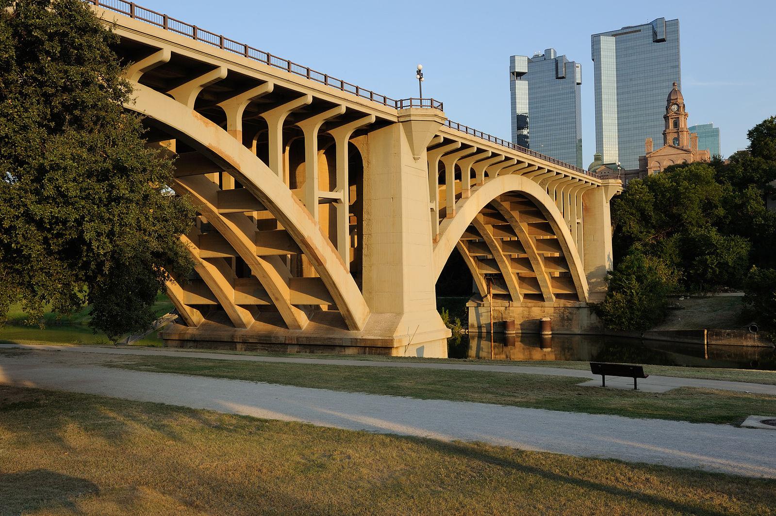 Grand concrete arch bridge over water with city buildings in golden light.