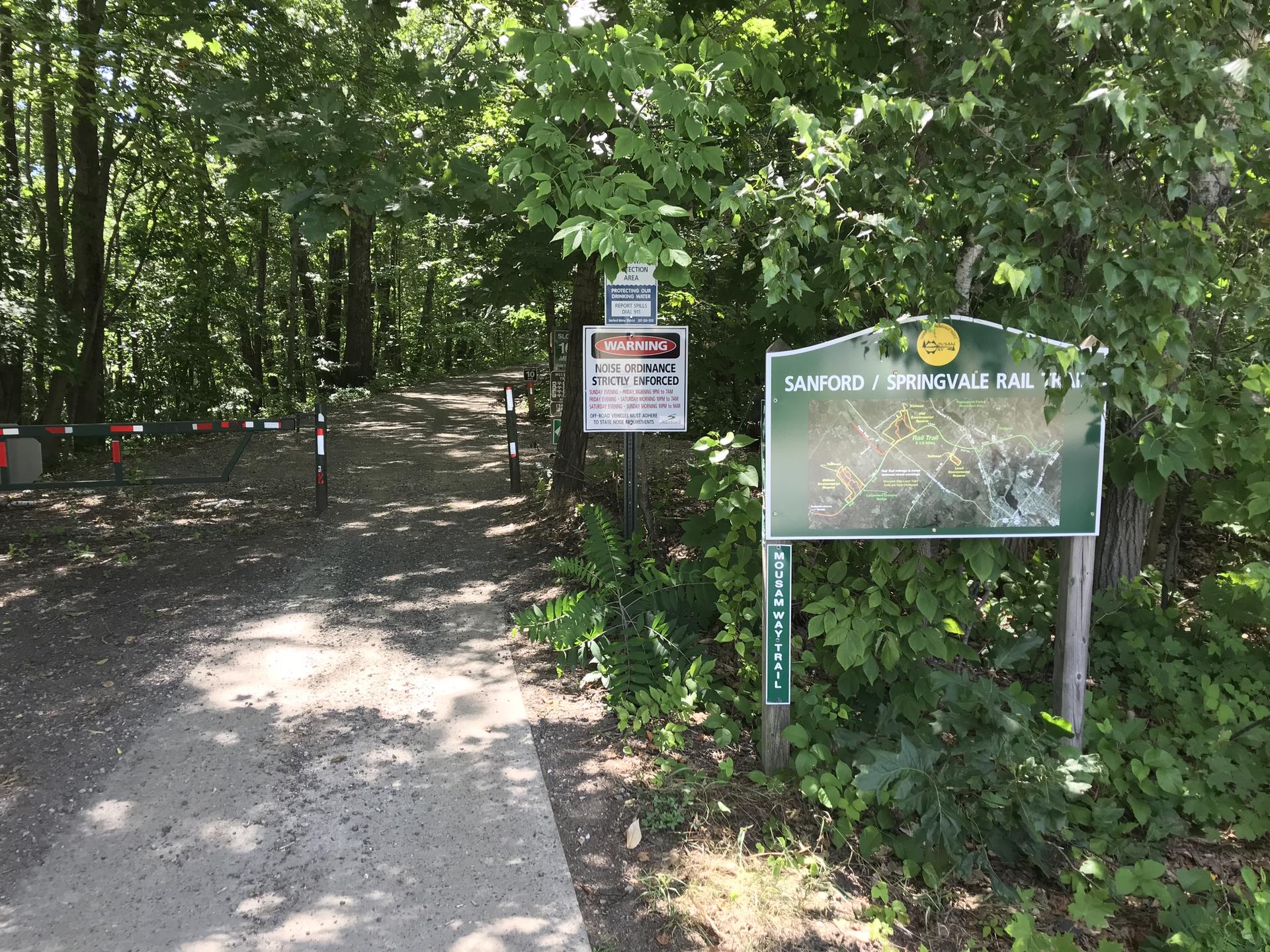 A shaded dirt path enters a dense forest, with signs and a barrier.