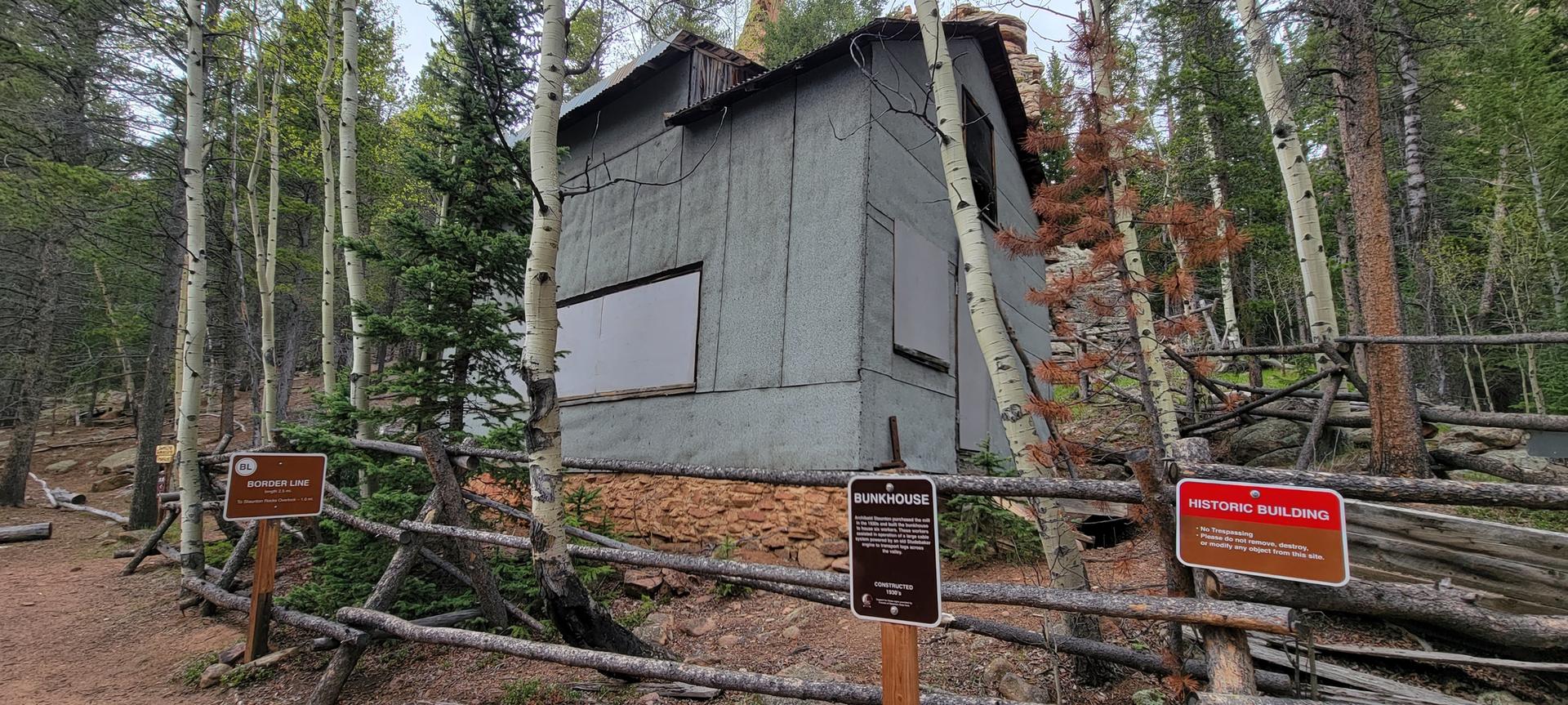 Weathered wooden building with boarded windows in a forest, surrounded by trees and a fence.