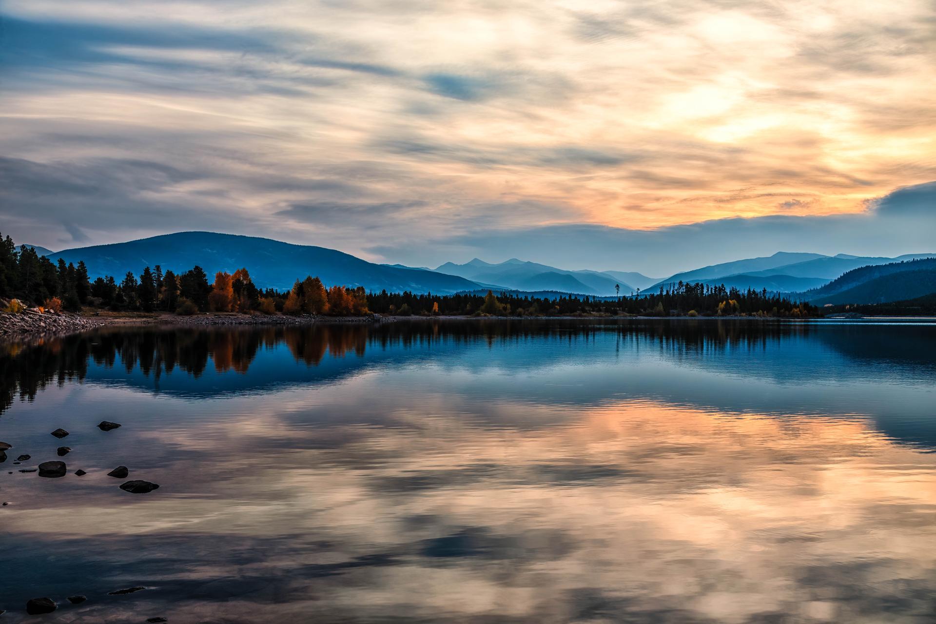 Calm lake reflecting a colorful cloudy sky and distant mountains at sunset.