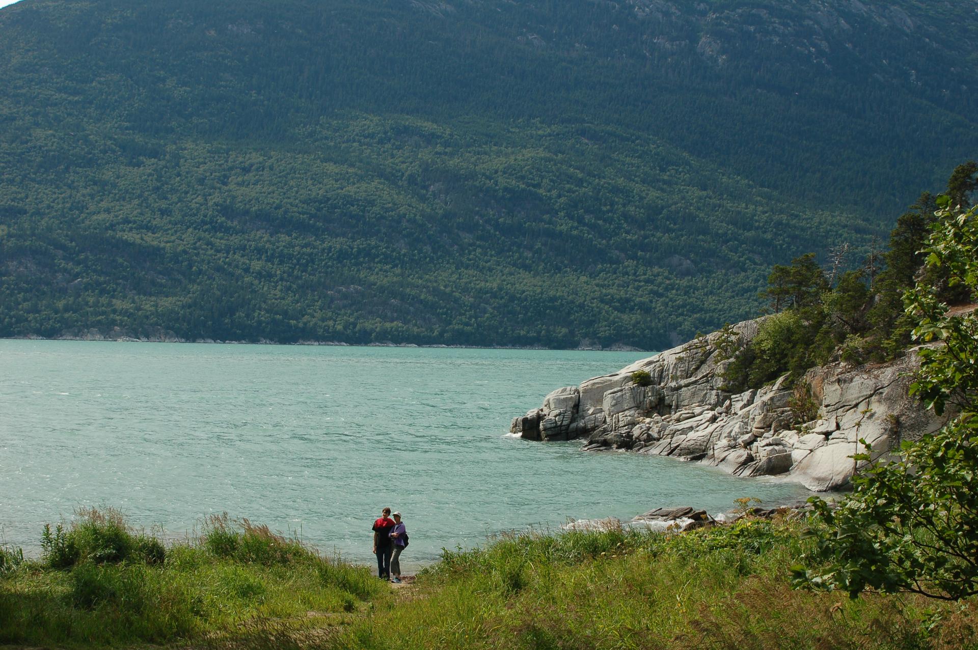 Two people stand by a calm lake with a large forested mountain.