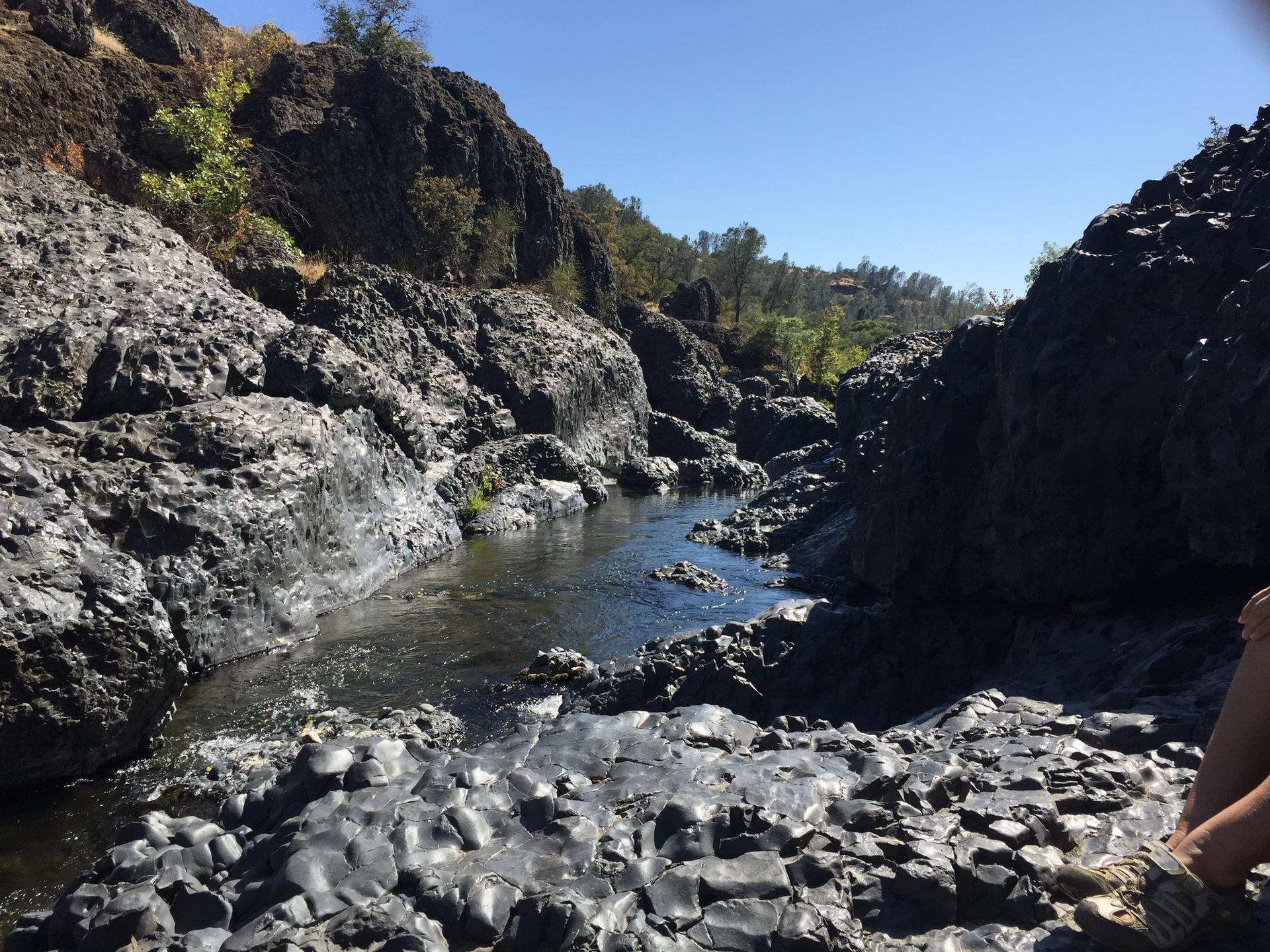 Narrow river flows through a rocky canyon under bright sun, person resting.