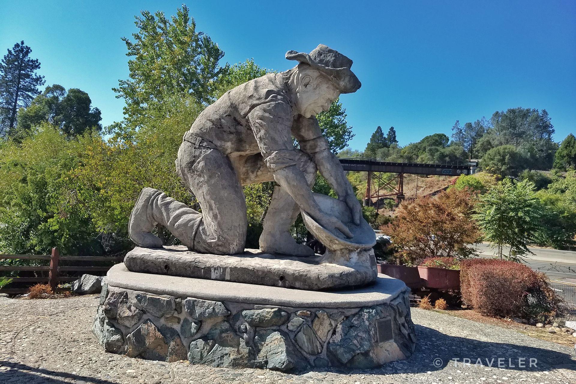 Stone statue of a person panning for gold under a bright blue sky.