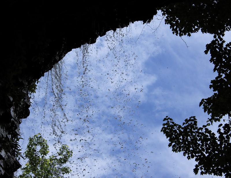 Water cascades from a dark rock overhang against a bright blue sky.