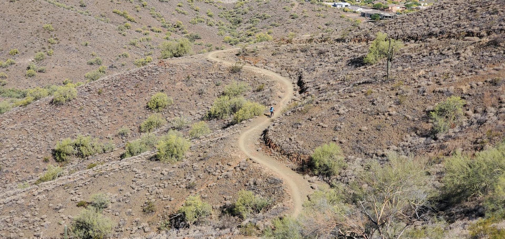 Winding dirt trail on a dry, rocky hillside under bright sunlight.