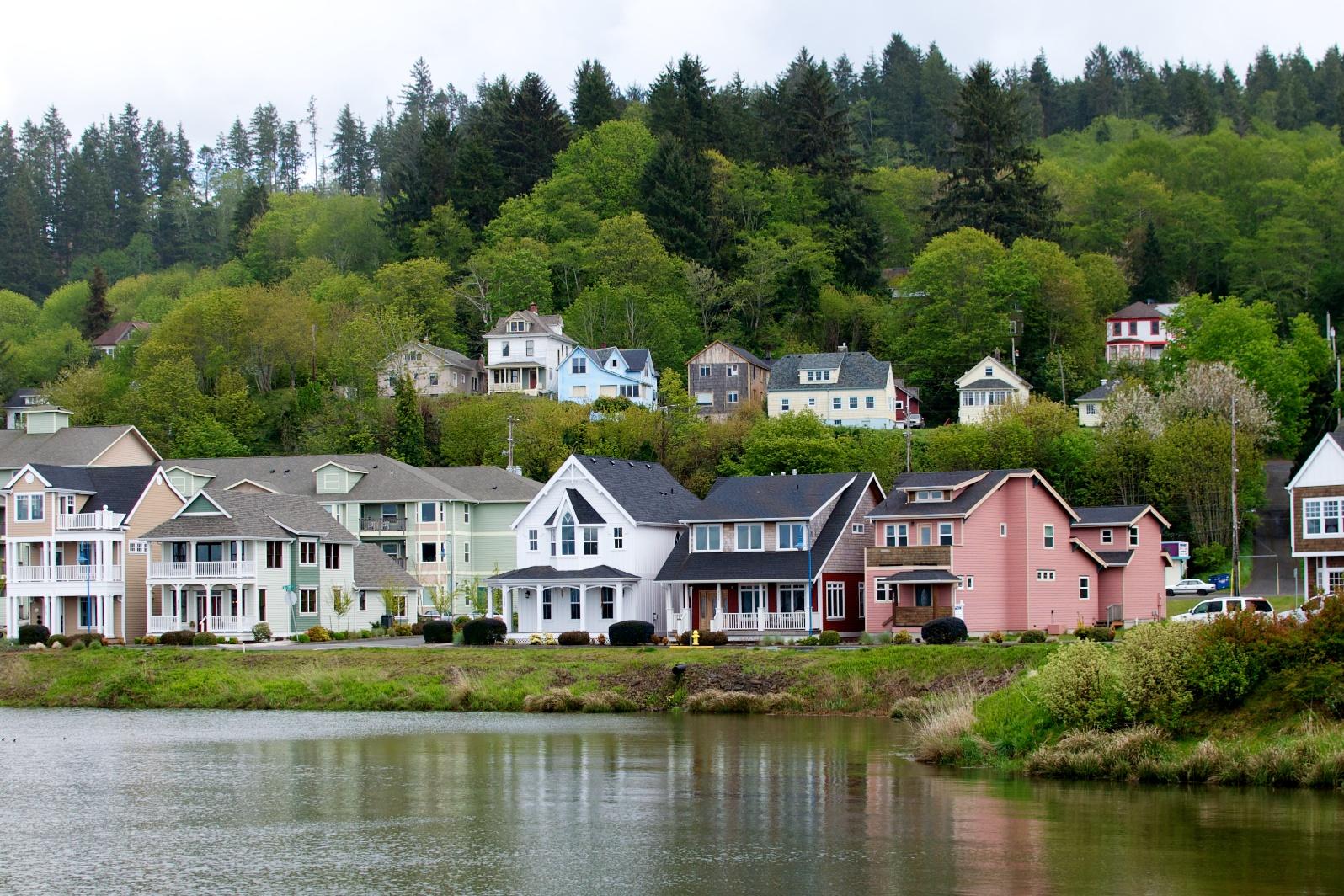 Waterfront houses with a steep, tree-covered hill under an overcast sky.