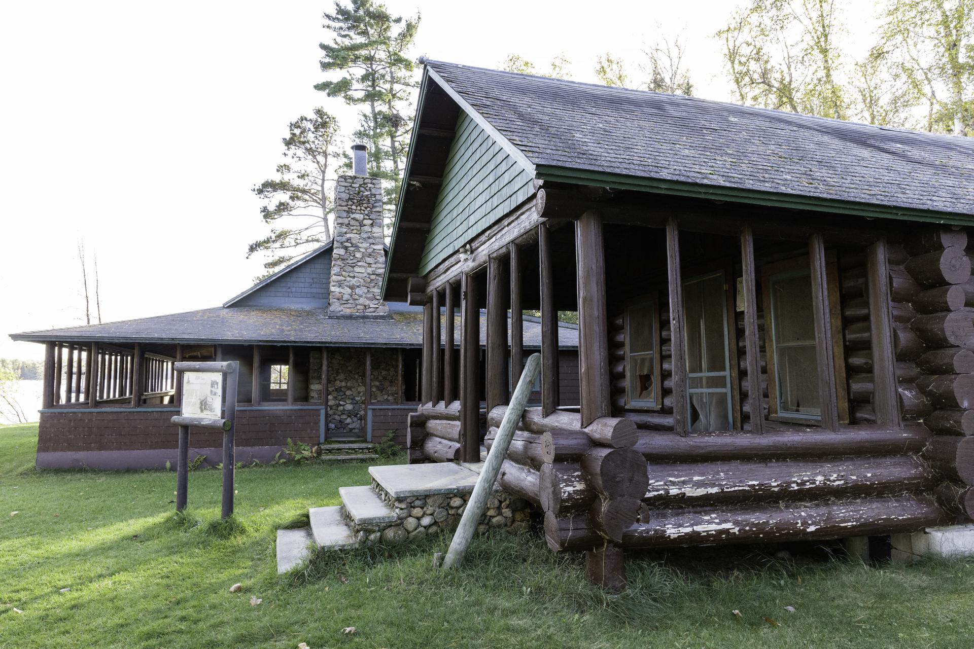 Rustic log cabin with a stone chimney and porch, surrounded by grass.