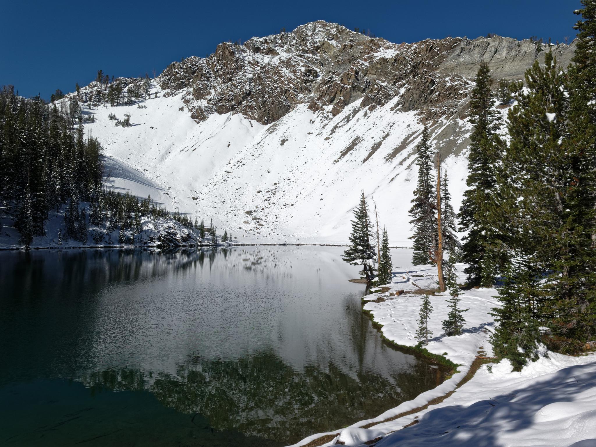 Snow-covered mountains reflect in a calm lake, surrounded by pine trees.