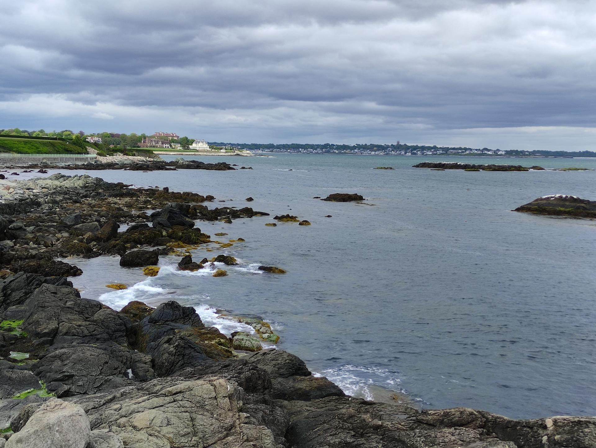 Overcast view of a rocky coastline with waves and distant buildings.