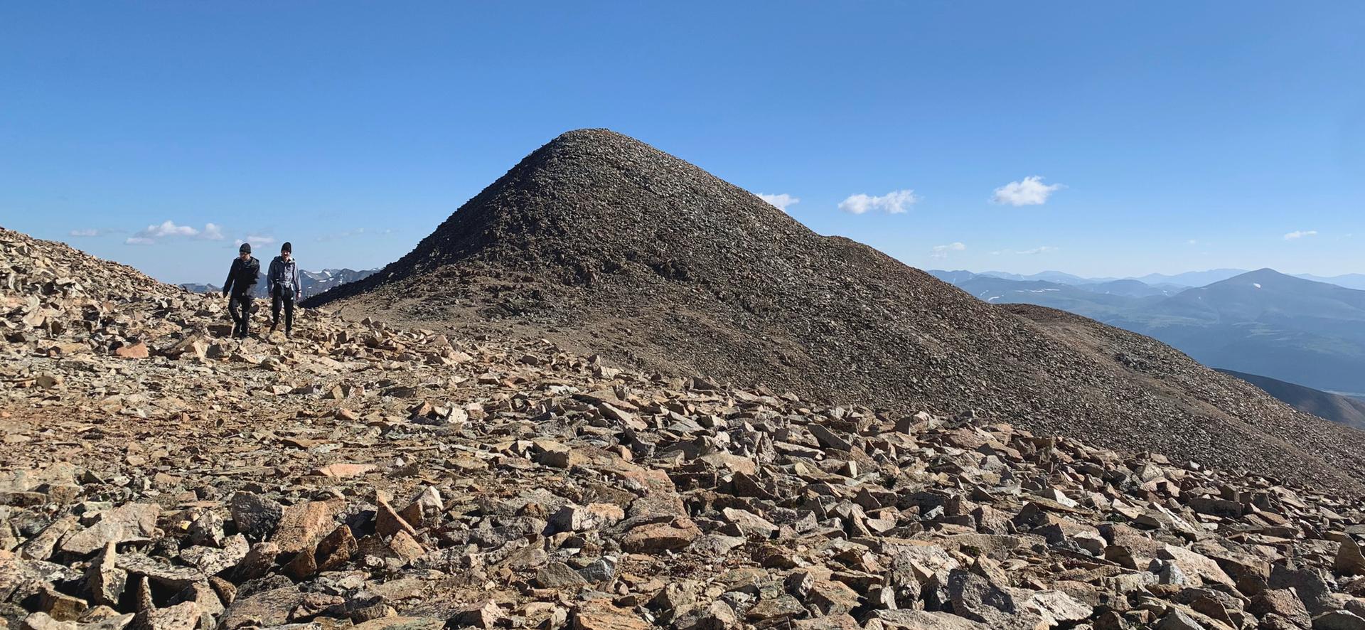 Two hikers walk on a rocky mountain path towards a distant peak.
