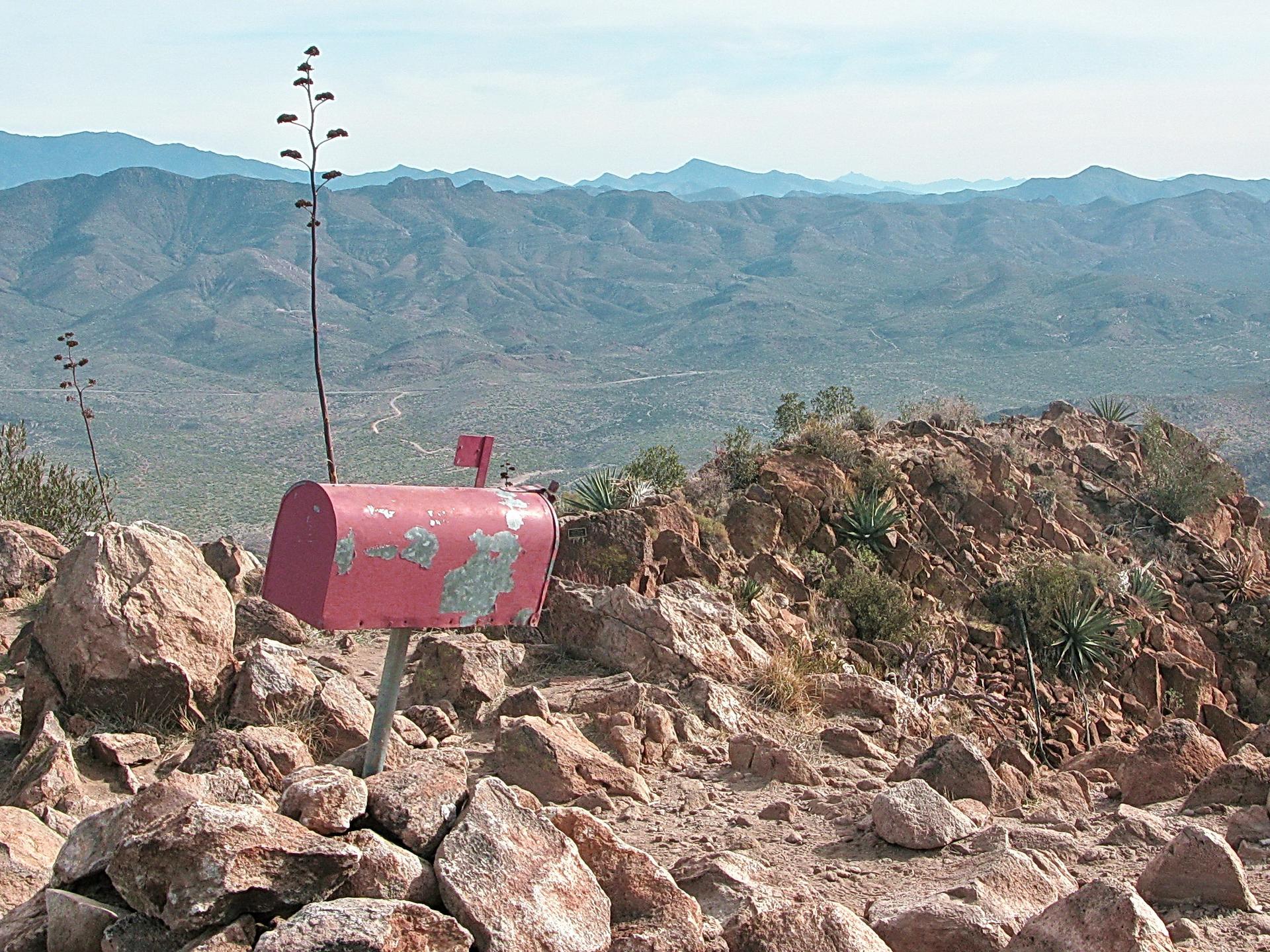 Weathered red mailbox on a rocky desert hilltop with distant mountains under bright sky.