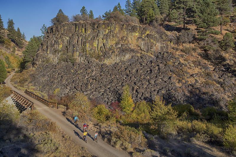 Three cyclists on a dirt path beside a large rocky cliff and forest.