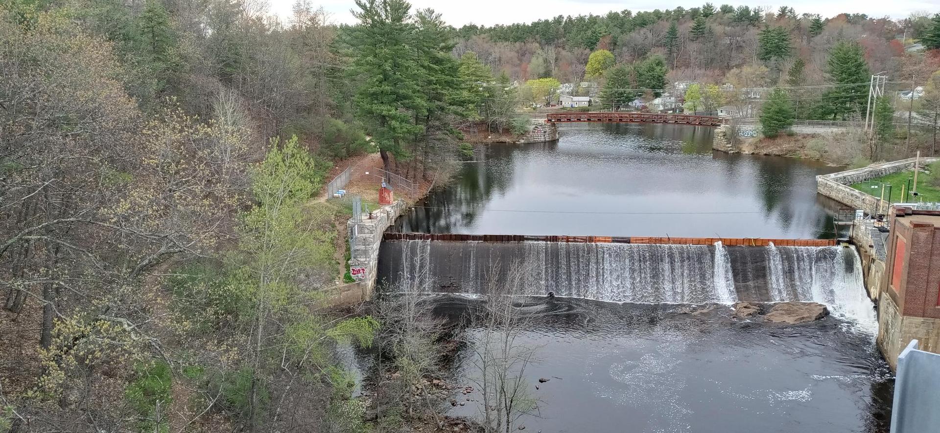 River with a dam and waterfall, surrounded by trees and distant bridges.