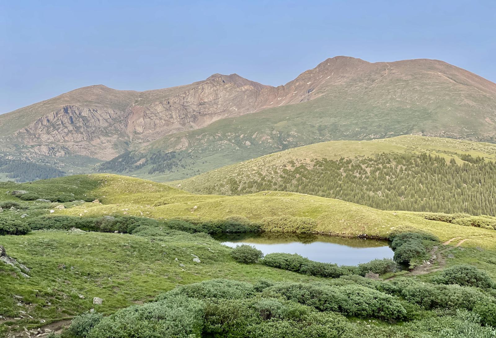 Small pond nestled in green hills with large mountains under soft natural light.