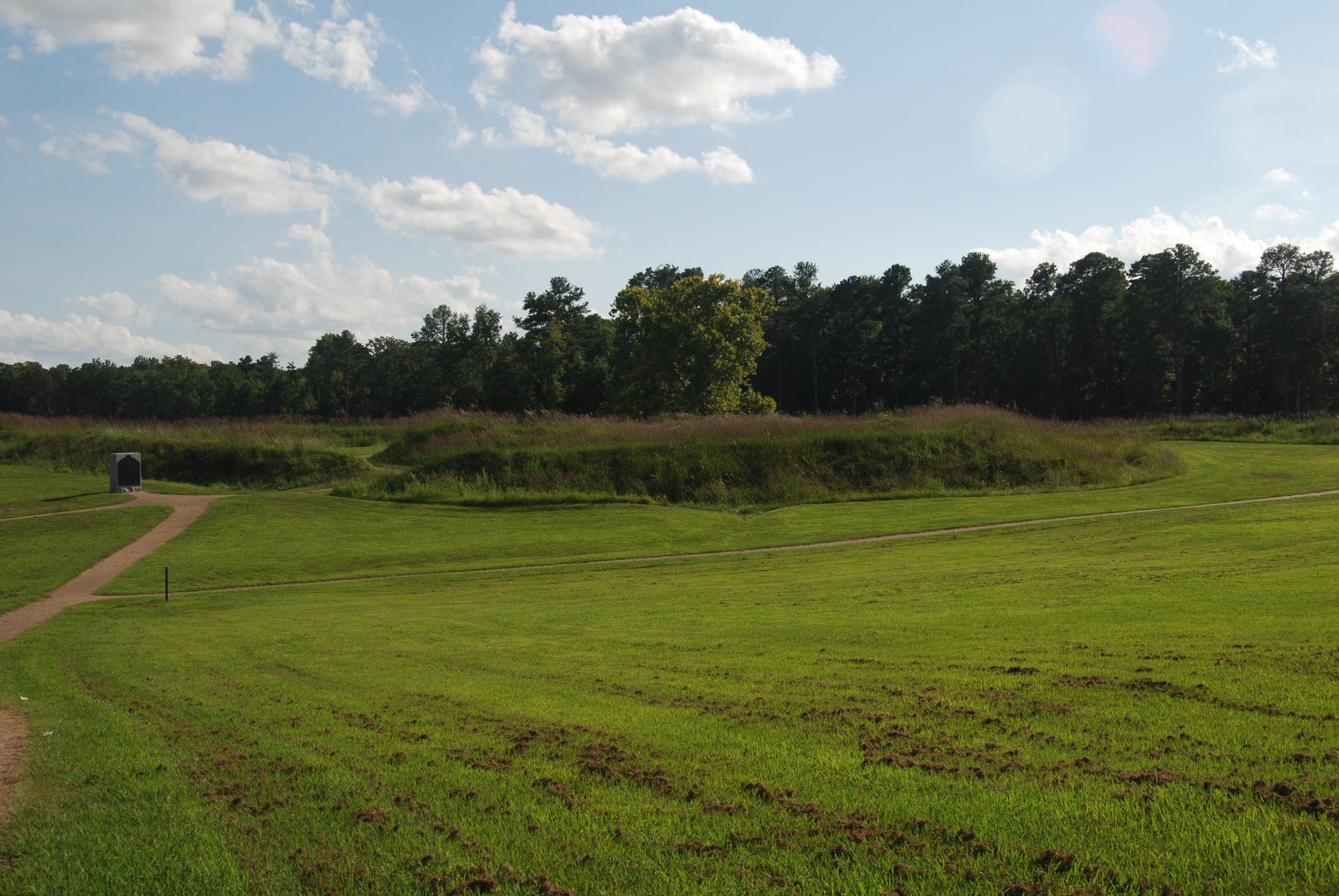 Expansive green field with rolling earthen mounds under a bright, cloudy sky.