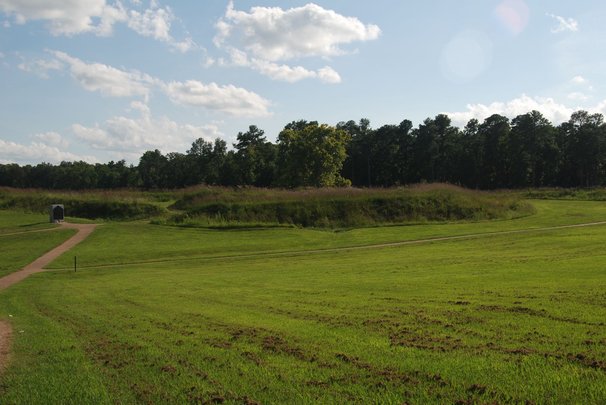 Expansive green field with rolling earthen mounds under a bright, cloudy sky. - Encampment Loop Trail