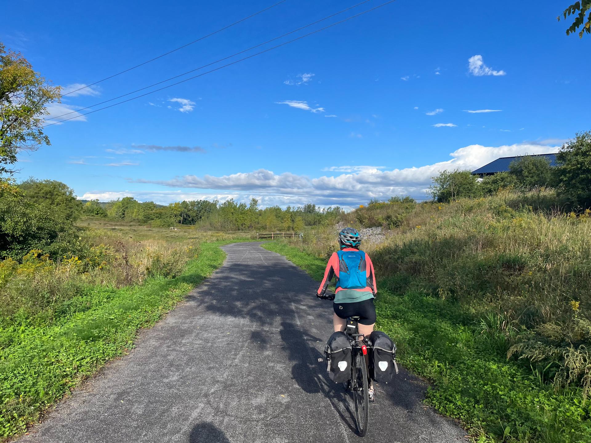 A person cycles away on a paved path through a sunny, grassy, treed landscape.