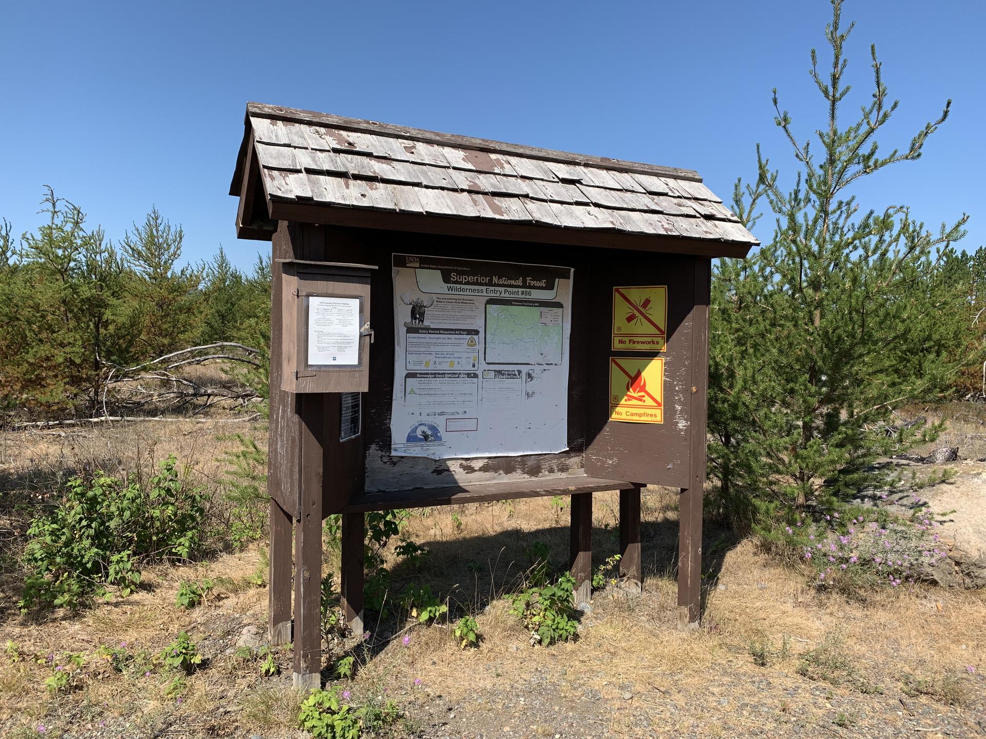 A wooden information kiosk stands in a dry, sunny field with pine trees.