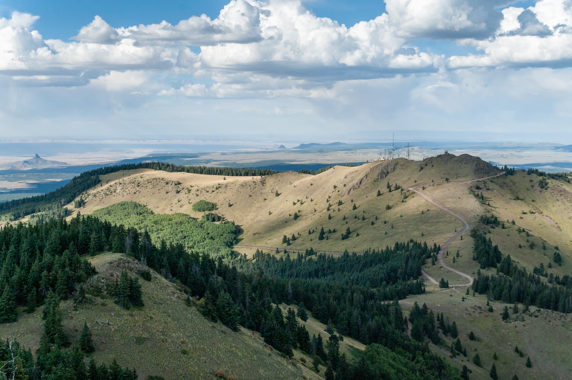 Expansive mountain landscape with green forests, dirt roads, and distant plains.