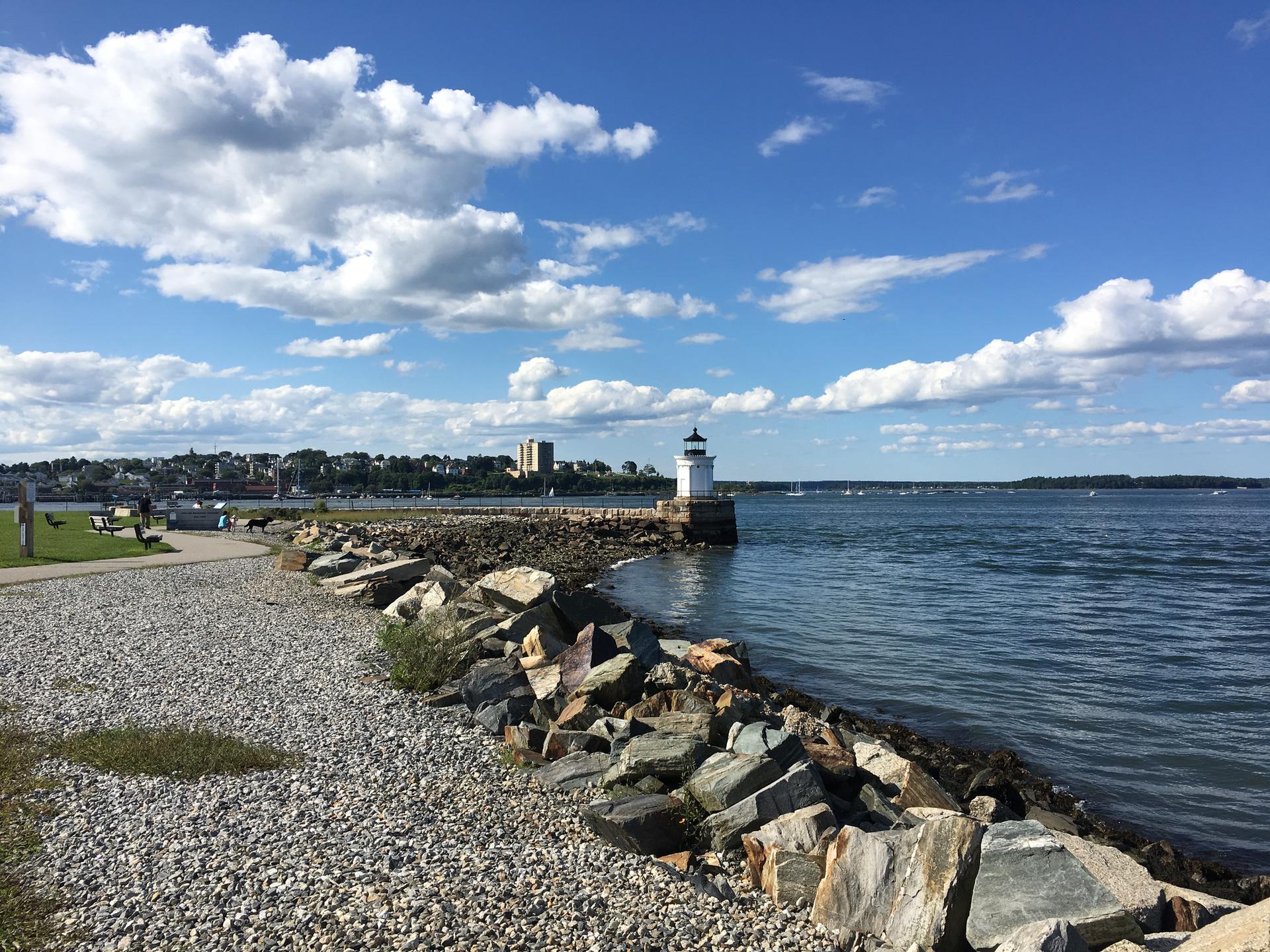 Sunny day, white lighthouse on a rocky jetty with a distant city.
