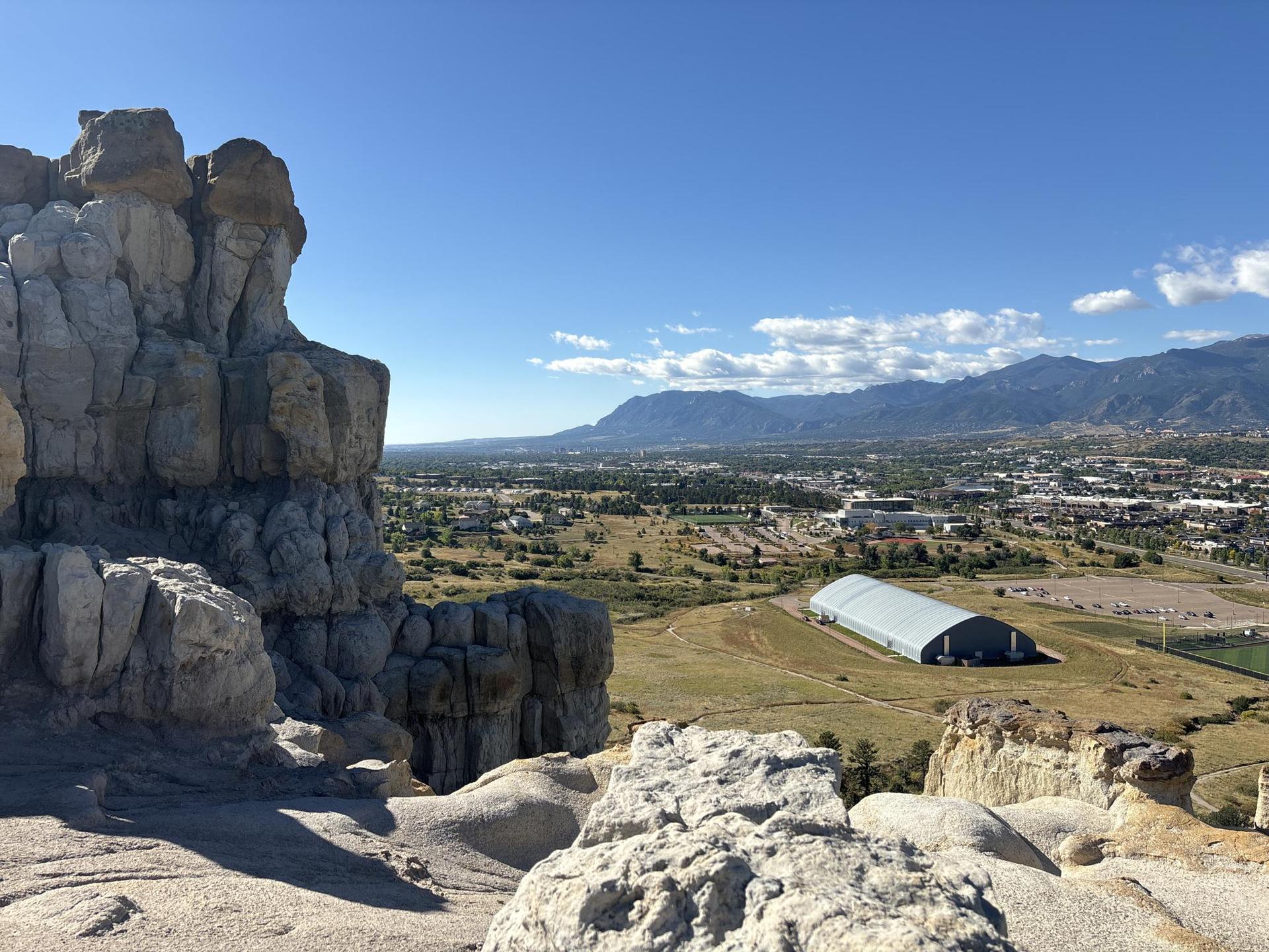 Large rock formations overlook a city and distant mountains under a clear sky.