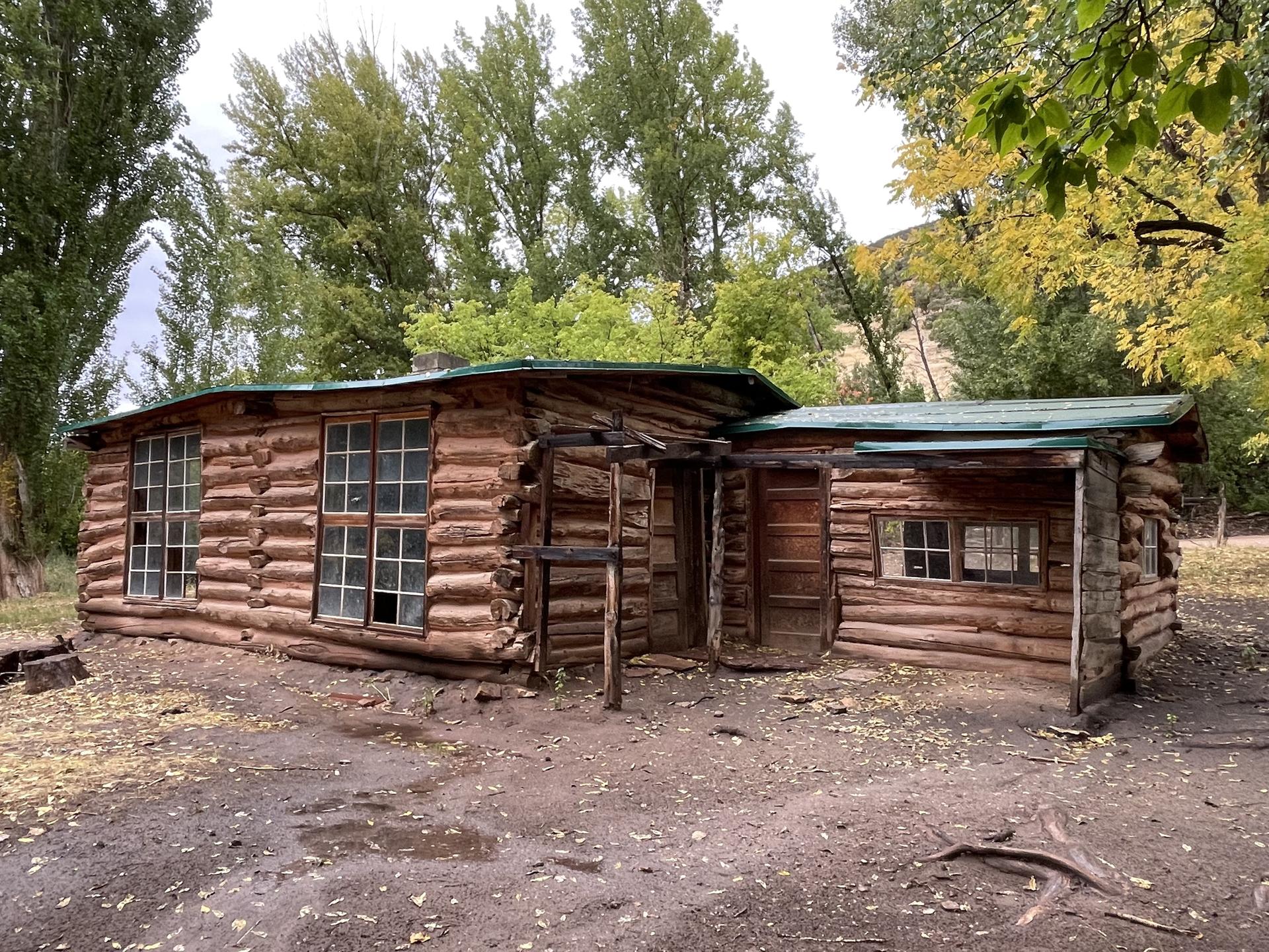 Old wooden log cabin with multiple windows, surrounded by trees.