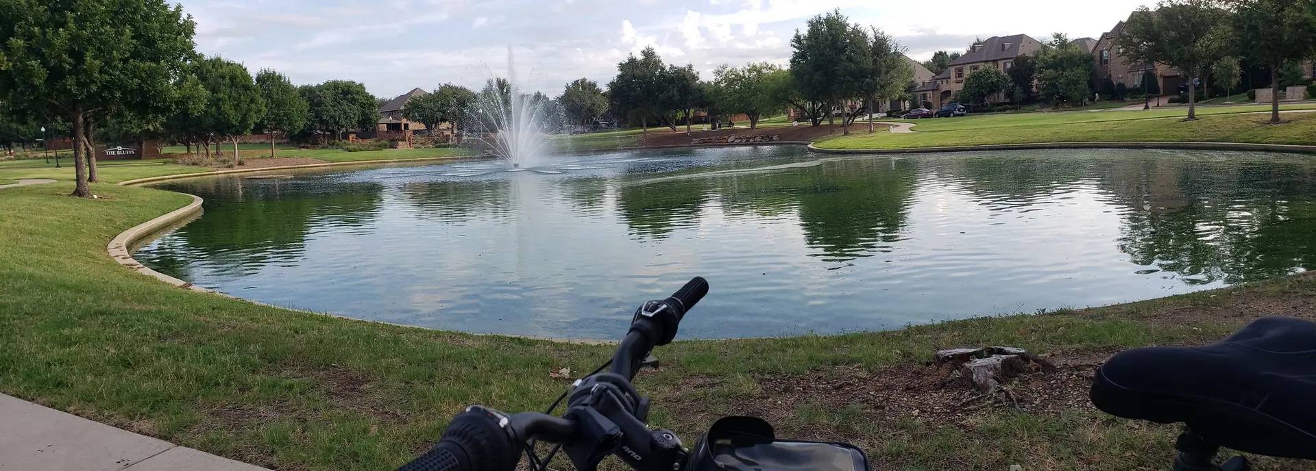 Bicycle view of a park pond with a fountain under an overcast sky.