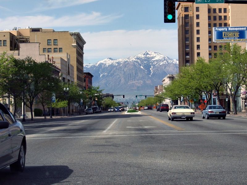 City street with cars, buildings, and snow-capped mountains in bright daylight. - Pineview Reservoir via North Ogden Canyon Road loop