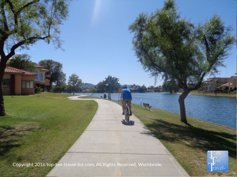 A person cycles on a sunny paved path beside a calm lake. - Camelback Walk Greenbelt – McDowell Sonoran Preserve loop from Chaparral High School